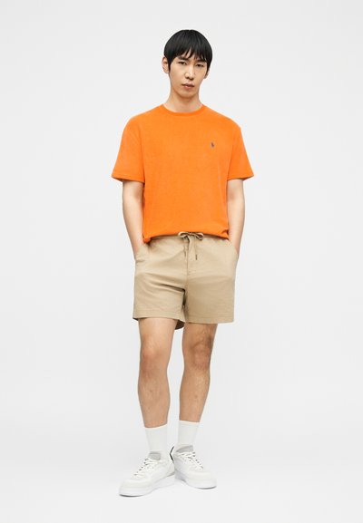 Άνδρας όρθιος, φοράει πορτοκαλί κοντομάνικο πουκάμισο, μπεζ σορτς με κορδόνι, λευκές κάλτσες και λευκά αθλητικά παπούτσια, μπροστά σε απλό λευκό φόντο.