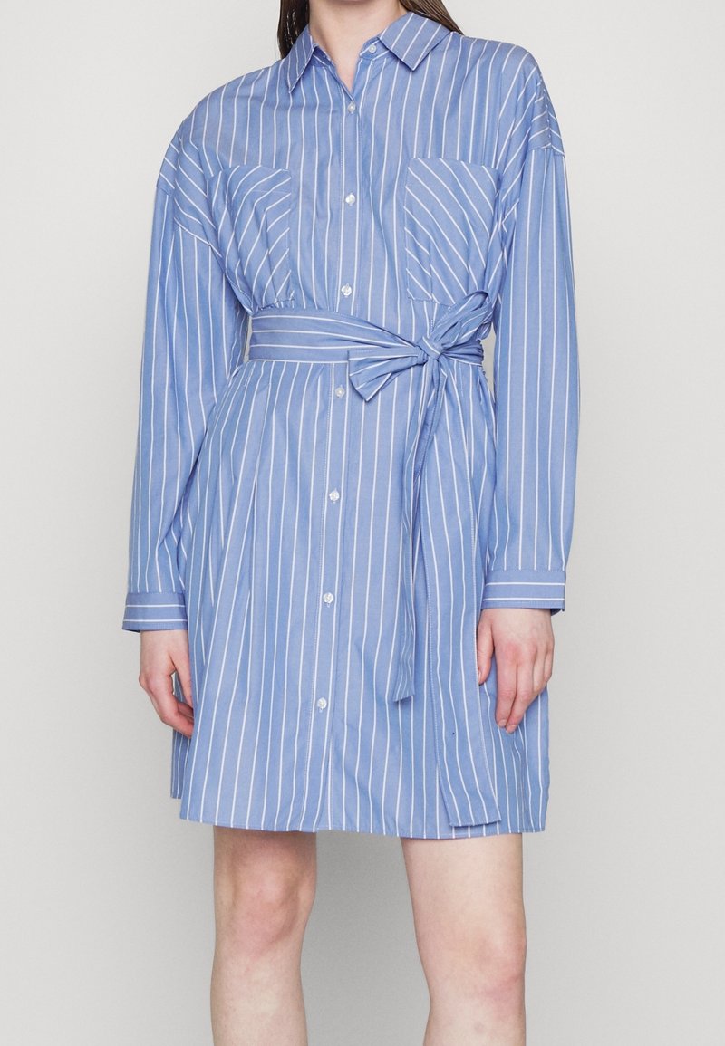 Robe chemise à rayures bleues avec manches longues, devant boutonné, deux poches poitrine, une ceinture nouée à la taille et des rayures horizontales.