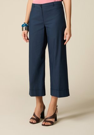 Donna che indossa pantaloni navy a gamba larga e corti, sandali con tacco marroni, top rosa e braccialetti spessi sul polso sinistro.
