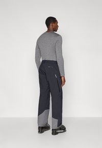 Γκρι μακρυμάνικο πουκάμισο φορεμένο με σκούρο μπλε παντελόνι σκι που διαθέτει γκρι πάνελ και τσέπες με φερμουάρ· το μοντέλο εμφανίζεται από πίσω.