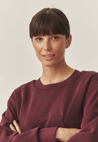 Bordeaux sweatshirt met lange mouwen, geribbelde ronde halslijn en een zachte textuur. Het model heeft de armen over elkaar, waardoor de pasvorm van het kledingstuk goed te zien is.