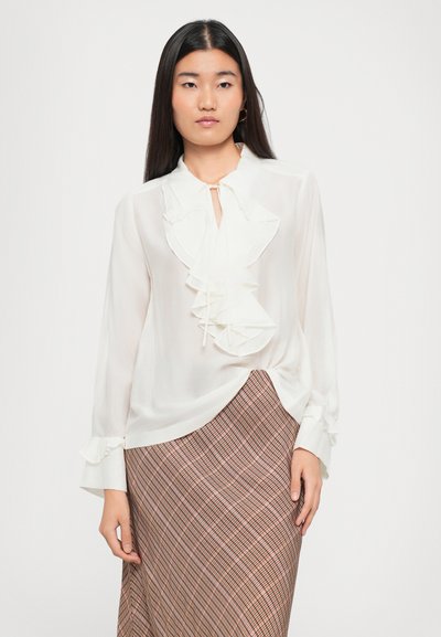 White, sheer blouse featuring ruffled detail down the front and a keyhole neck. Long sleeves with ruffle cuffs. Paired with a checked skirt.