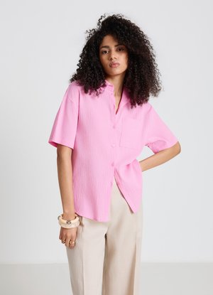 Woman with curly hair wearing a loose pink short-sleeve button-up shirt and beige pants, standing with one hand on hip.
