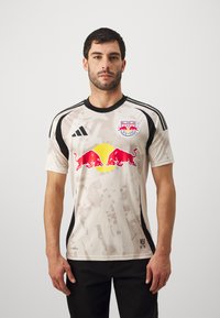 Světle šedý fotbalový dres s maskovacím vzorem, černými akcenty a krátkými rukávy. Obsahuje logo Red Bull, žlutý kruh a značku Adidas.