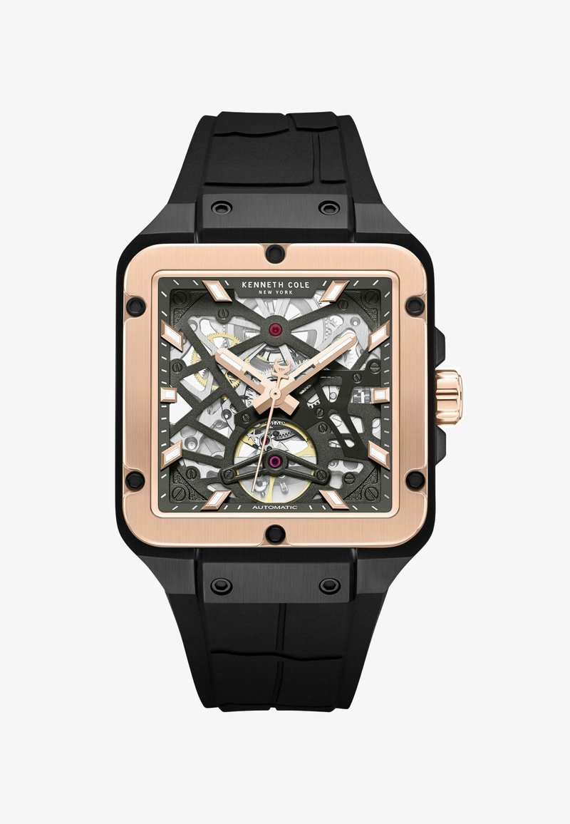 Montre carrée avec un bracelet en caoutchouc noir, verre transparent révélant les rouages, accents dorés et détails roses sur le cadran.
