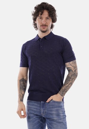 Uomo con capelli ricci che indossa una polo blu navy dall'aspetto strutturato e jeans blu, mostrando tatuaggi dettagliati su entrambi gli avambracci.