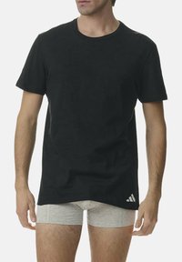 T-shirt en coton noir à manches courtes, col rond, avec un logo adidas blanc en bas à gauche. Porté par-dessus des boxers gris clair.