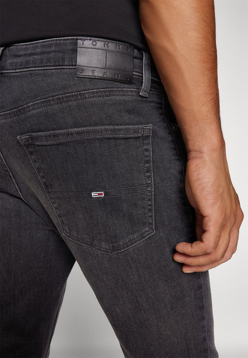 Vaqueros de mezclilla gris oscuro con un parche de cuero que lleva la marca "Tommy Jeans", un bolsillo trasero con un pequeño logo y un diseño ajustado.