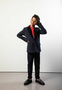 Granatowa marynarka garniturowa z czarnymi klapami, czerwony golf i granatowe spodnie. Do kompletu czarne mokasyny. Prosta, gładka tekstura, sylwetka całej postaci.