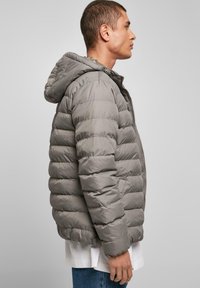 Grå pufferjakke med hætte, prydet med vandret quiltet design, lynlås lukning og to sidelommer. Båret over en hvid skjorte.