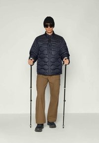 Veste d'hiver en duvet navy avec un design matelassé, pantalons marron et bottes de randonnée noires. Le mannequin tient des bâtons de trekking, portant un bonnet et des lunettes de soleil.