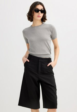 Mujer con pantalones cortos negros de pierna ancha, camiseta gris de manga corta y gafas de sol negras, de pie con las manos en los bolsillos sobre fondo blanco.