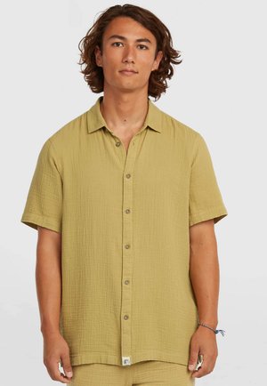 Noor mees keskmise pikkusega pruuni juustega, kandes lõdva istuvusega lühikeste varrukatega kollakas-kollast nööbitavat särki.