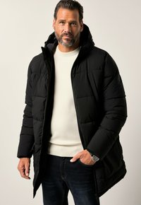 Schwarze Puffjacke mit Kapuze, gestepptem Design, Frontreißverschluss und seitlichen Taschen. Tragen über einem hellen Pullover und dunklen Jeans.