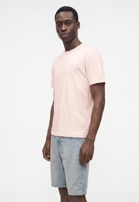 Άνδρας που φοράει ροζ ανοιχτό κοντομάνικο T-shirt και ανοιχτό μπλε τζιν σορτς με κεντημένο αστέρι στο αριστερό πόδι, στέκεται μπροστά σε μια μονόχρωμη φόντο.