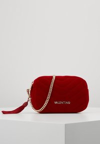 Czerwona aksamitna torebka z pikowanym wzorem, złotym łańcuszkiem i akcentami z frędzlami, o prostokątnym kształcie i wytłoczonym logo z przodu.