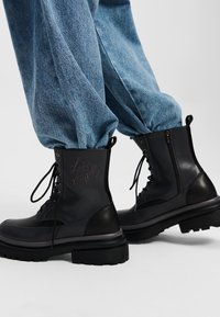Botas de combate de cuero negro con punteras redondas, suelas de goma estriadas, cordones en la parte delantera y cremallera lateral. Presentan letras en relieve en el cañón.