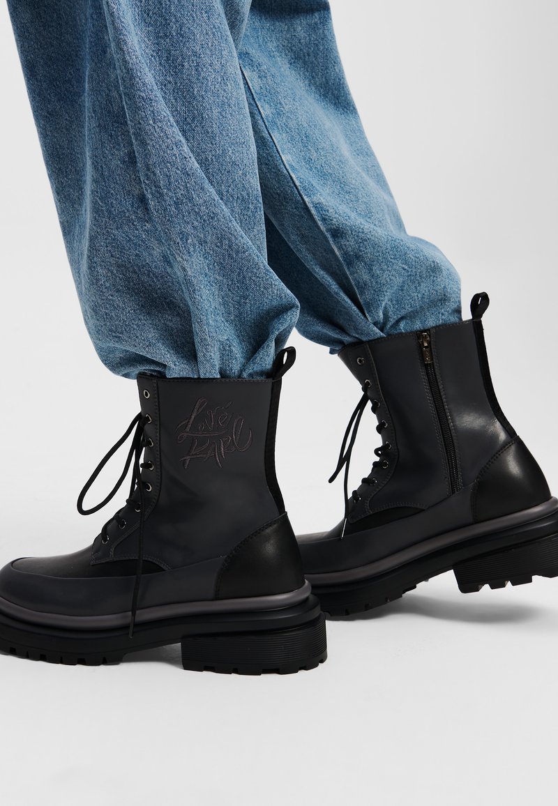 Botas de combate de cuero negro con punteras redondas, suelas de goma estriadas, cordones en la parte delantera y cremallera lateral. Presentan letras en relieve en el cañón.
