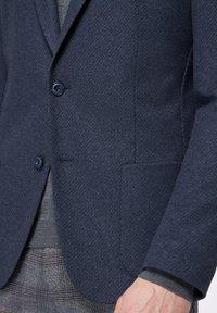 Blazer blu navy con texture a spina di pesce, due bottoni e una tasca. Indossato sopra un maglione grigio con polsino visibile sotto la manica.