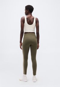 Γυναίκα που στέκεται πλάτη, φοράει λευκό κοντό τοπ, χακί κολάν με πλευρικές ρίγες, λευκές κάλτσες και λευκά αθλητικά παπούτσια.