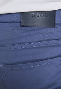 Pantalones de tela azul con una textura suave, detalles de costura y un parche de cuero negro con el logotipo "BOSS" en relieve en la parte posterior de la cintura.
