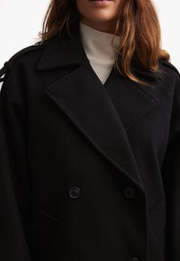 Cappotto doppiopetto in misto lana nera con ampi risvolti, caratterizzato da bottoni neri e tasche laterali, indossato sopra un maglione girocollo crema.