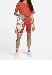 Kvinna iklädd röda Nike cykelbyxor och T-shirt, håller en röd och vit tie-dye sweatshirt, vita sneakers och rosa strumpor.