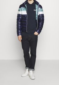 Mężczyzna ubrany w granatową, turkusową i białą kurtkę puchową w paski, czarną koszulę polo, ciemne dżinsy z mankietami i białe sneakersy, stoi z jedną ręką w kieszeni.