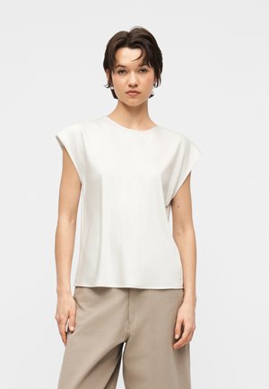 Ung person med kort mørkt hår iført en hvid ærmeløs top og beige bukser med vide ben, stående mod en ensfarvet baggrund.