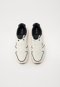 White sneakers with textured material, black accents on the collar and sole, lace-up design, featuring a subtle logo and multi-colored stripes.
