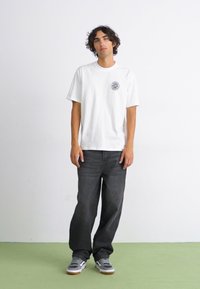 Hvid bomulds t-shirt med et rundt logo på venstre bryst, kombineret med mørkegrå jeans og grå sneakers med hvide og brune detaljer.
