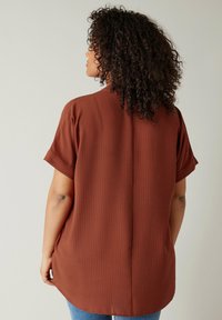 Camisa marrón de manga corta con un patrón texturizado, corte relajado y dobladillo curvado. Vista trasera destacada por un ligero detalle en el cuello.