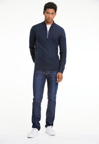 Merinovillapaita, jossa on puolen vetoketjun kaulus, yhdistettynä tummansinihin farkkuihin ja valkoisiin lenkkareihin. Tasainen pinta, istuva malli, vähäiset korostukset.