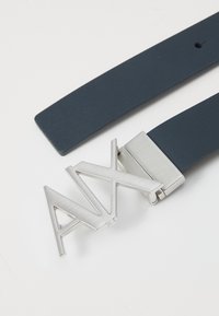 Mørk marineblå syntetisk lærbelte med en sølvfarget metallspenne som har vinklet, utskåret bokstaver som staver "ANX." Glatt tekstur med minimalistisk design.