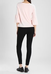 Světle růžový top s rozšířenými rukávy, kombinovaný s přiléhavými černými kalhotami a černými botami, stojící na neutrálním pozadí.