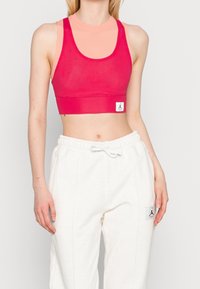 Pink mesh sports crop top with wide straps, layered over a light peach base layer. Paired with white jogger pants featuring logo patch.