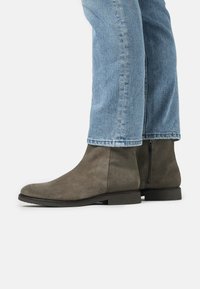 Botas de tobillo de ante gris con una textura suave, cremallera lateral y suela plana de goma negra, combinadas con pantalones de mezclilla azul claro.