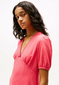 Vestido rosa con escote en V, mangas abullonadas y un detalle de lazo en el frente. Presenta una textura suave y acentos fruncidos en los dobladillos de las mangas.
