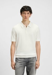 Kortærmet polo skjorte, hvid, tekstureret stof, tre-knaps placket, ribbede manchetter, båret med lysegrå slidte jeans.