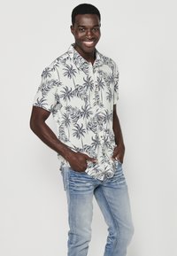Camicia bianca a maniche corte con stampa di foglie di palma nera, design con bottoni, abbinata a jeans blu chiaro. Modello in posa con le mani nelle tasche.