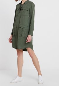 Olivově zelené šaty s dlouhým rukávem, límečkem, dvěma náprsními kapsami, stahovacím detailem a mírně asymetrickým lemem; v kombinaci s bílými teniskami.
