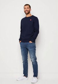 Laivastonsininen collegepaita pyöreällä kaula-aukolla ja punaisella logolla rinnassa, yhdistetty vaaleansinisiin farkkuihin ja valkoisiin lenkkareihin.