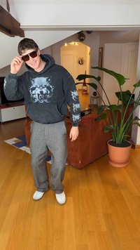 Must kapuutsiga dressipluus hundi graafikaga, halli mustriga püksid ja valged tossud; kannab tumedaid päikeseprille, seisab toas taime kõrval.