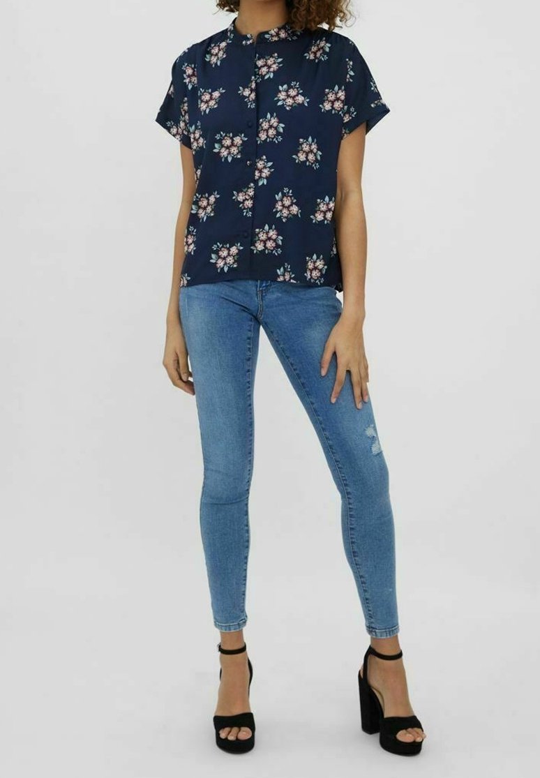 Chemise fleurie bleu marine à manches courtes, avec des boutons sur le devant, portée avec un jean skinny bleu clair. Sandales noires à talons hauts complètent la tenue.