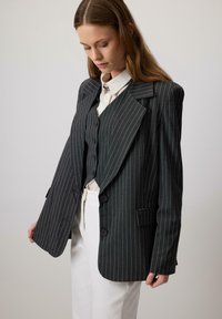 Vrouw in een oversized donkergestreepte blazer en vest over een wit shirt en broek, kijkt naar beneden en houdt de rand van de blazer vast.