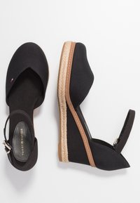 Paire d'espadrilles compensées noires avec brides à la cheville et détail tissé sur la semelle, une chaussure vue de dessus, l'autre de côté.