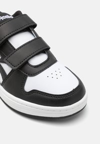 Chaussure de sport noir et blanc avec une tige en cuir lisse, des bandes Velcro, une pointe perforée et une semelle en caoutchouc plate.