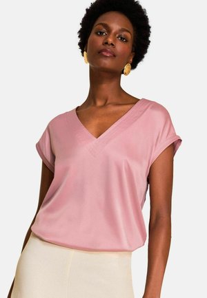 Kobieta ubrana w jasnoróżowy top z dekoltem w serek i krótkimi rękawami oraz kremowe spodnie, z dużymi złotymi okrągłymi kolczykami.