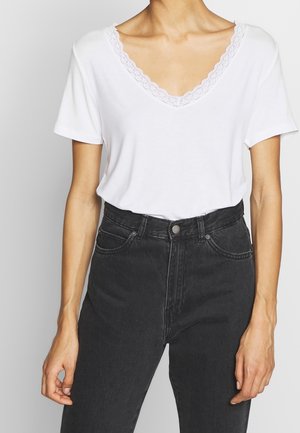 Kobieta ubrana w biały, krótki top z dekoltem w serek i koronkowym wykończeniem, włożony do wysokich czarnych jeansów, stojąca na tle jednolitego tła.