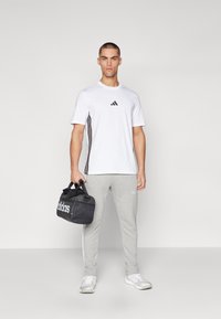 Witte sport T-shirt met zwart logo, grijze sweatpants met zijstrepen en zwarte sporttas. Model draagt lichte sneakers.